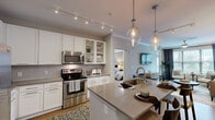 Located at 2875 Crescent Pkwy in Atlanta, GA, this well-appointed two-bedroom, two-bathroom residence offers a thoughtfully designed single-level layout. The open-concept kitchen and living area serves as the heart of the home, featuring crisp white cabinetry, a subway tile backsplash, stainless steel appliances including a refrigerator with ice maker, electric range, and built-in microwave. A large center island with a quartz countertop and undermount sink provides additional prep space and seating, illuminated by elegant glass pendant lights and a track lighting system overhead. Warm hardwood-style flooring flows throughout the main living areas. The living room is bright and inviting, with large windows framing outdoor greenery and a glass-paneled door that fills the space with natural light. Both bedrooms are generously proportioned with plush carpet, ceiling fans, and windows with outdoor views. The primary bedroom offers a comfortable retreat with dedicated closet space. Each bathroom is finished with neutral tile, vanities with quartz counters, and a tub-shower combination. Two closets and a connecting hallway complete the functional layout of this modern Atlanta residence.
