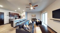 This single-story space showcases a thoughtfully designed open-concept layout that seamlessly connects the kitchen, dining, and living areas into one cohesive environment. The kitchen is outfitted with dark espresso cabinetry, stainless steel appliances including a range and microwave, and light granite countertops that extend into a generous breakfast bar with tall bar-height seating — ideal for casual dining or entertaining. Recessed lighting throughout the kitchen ensures the workspace is well-lit and functional.

The living area flows naturally from the kitchen and features warm hardwood-style flooring that runs throughout the space, adding continuity and a sense of warmth. A large sectional sofa anchors the room, complemented by accent chairs and a low-profile cube storage unit used for display. A ceiling fan with a wood-tone blade finish is centrally mounted, providing both airflow and ambient light.

Natural light enters generously through a sliding glass door on the far wall, which opens to what appears to be a covered outdoor area. The neutral gray wall tones and modern furnishings throughout contribute to a clean, contemporary aesthetic that feels both comfortable and practical.