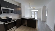This single-floor space showcases a thoughtfully designed open-concept layout that seamlessly connects the kitchen and living areas. The kitchen is outfitted with dark espresso cabinetry that contrasts sharply against white subway tile backsplash and light grey stone countertops, creating a modern, high-contrast aesthetic. Stainless steel appliances, including a range, built-in microwave, and dishwasher, are integrated throughout. The peninsula-style counter extension features an undermount sink and provides additional workspace while acting as a natural divider between the kitchen and the adjoining living area. Light wood-look plank flooring runs throughout, adding warmth and continuity to the space. A multi-bulb pendant light fixture hangs above the peninsula, offering focused task lighting. The living area benefits from generous natural light streaming through a large sliding glass door or French door that opens to an outdoor balcony or patio, enhancing the sense of openness. White walls and ceilings keep the interior feeling bright and airy, complementing the clean, contemporary design throughout.