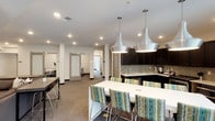 This single-floor amenity space is thoughtfully designed to serve as a multifunctional gathering area, blending a fully equipped kitchen, open lounge, and semi-private rooms into one cohesive layout. The kitchen is a standout feature, outfitted with dark espresso cabinetry extending to the ceiling, a mosaic tile backsplash, stainless steel appliances including a range, microwave, and French door refrigerator, and bright white countertops. A large central island with striped upholstered bar seating and striking metallic pendant lights overhead adds both function and visual interest. The flooring transitions from large-format hexagonal tile in the kitchen to plush carpet throughout the lounge and corridor areas. The lounge is anchored by comfortable sectional seating, a wall-mounted flat-screen television, and a bold accent wall featuring dark paneling with yellow horizontal stripes. Recessed lighting throughout provides consistent illumination. Frosted glass barn doors line a corridor leading to smaller private rooms, offering flexible space for meetings or individual use. A secondary lounge area with direct access to an outdoor patio allows natural light to filter in, enhancing the overall ambiance of the space.