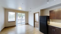 This single-floor residence offers a clean, modern design with a warm and functional layout. The open-concept living area is finished with light hardwood-style flooring that extends throughout, creating a bright and cohesive atmosphere. Neutral-toned walls complement the natural light that flows in generously through a large sliding glass door, which opens onto a private balcony with a metal railing and pleasant outdoor views of surrounding trees. The kitchen is equipped with a full-size black refrigerator and connects seamlessly to the living space, making it ideal for everyday living and entertaining. A doorway from the main living area leads to an adjoining carpeted room, offering a comfortable and distinct space that serves as a bedroom or additional retreat. The unit features flush-mounted ceiling lighting, visible HVAC venting, and a smoke detector, reflecting thoughtful and practical construction throughout. The overall ambiance is clean, understated, and move-in ready, with a simple aesthetic that provides a versatile backdrop for a variety of interior styles.