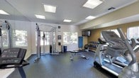 This fitness center offers a comprehensive workout environment designed to support a variety of exercise routines. The space is finished with durable rubber flooring throughout, providing traction and cushioning ideal for both strength training and cardio activities. Overhead, large recessed panel lights illuminate the room evenly, creating a bright and energizing atmosphere.

On the strength training side, the room features a multi-function cable weight machine, a flat utility bench, and a well-stocked dumbbell rack offering a range of free weights. A stability ball and additional accessories are also present for versatile training options. For cardio, at least one treadmill and an elliptical machine are visible, positioned for convenient use.

A wall-mounted flat-screen television is installed at an elevated position, allowing users to watch content during their workouts. Windows along one wall let in natural light and provide views of the surrounding outdoor greenery, contributing to an open and pleasant ambiance. The neutral-toned walls and clean layout give the space a polished, functional feel. Overall, this fitness center is thoughtfully organized to maximize usability while maintaining a comfortable and motivating environment for residents or guests.