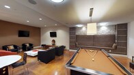 The Groveton Green amenity lounge is a thoughtfully designed, single-floor entertainment space that combines multiple recreational areas into one cohesive environment. The main lounge area features polished concrete flooring in warm amber tones throughout, recessed lighting, and a contemporary aesthetic defined by neutral tones with rich brown accent walls.

The billiards area anchors one end of the main space, equipped with a full-size pool table beneath a modern rectangular pendant light. Adjacent lounge seating consists of sleek black leather chairs and a white coffee table, arranged around a wall-mounted flat-screen TV and a built-in shelving unit with stone tile accents and a built-in bench.

A dedicated poker room offers an intimate setting with a branded Groveton Green round poker table, acrylic chairs, a decorative overhead pendant light, a wall-mounted TV, and built-in cabinetry with a granite countertop.

The wet bar area features dark cabinetry, stainless steel countertops and sink, a mosaic tile backsplash, and stylish pendant lighting. This area opens directly into a secondary seating lounge with additional leather chairs, a red accent armchair, and another wall-mounted TV, creating a versatile space well-suited for social gatherings.