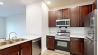 Located at 2100 Memorial in Houston's desirable 77007 corridor, this one-bedroom, one-bathroom residence offers a thoughtfully organized single-floor layout. The open kitchen and living area is anchored by a well-appointed kitchen featuring rich dark-stained cabinetry with brushed nickel hardware, marble-look quartz countertops, a classic white subway tile backsplash, and a full suite of stainless steel appliances including a range, over-the-range microwave, dishwasher, and double-basin sink. Recessed lighting brightens the space throughout. Light-toned hardwood flooring runs through the main living areas, complementing the clean, neutral color palette. The spacious primary bedroom features plush carpeting, a ceiling fan, and a large multi-pane window that frames outdoor greenery, filling the room with natural light. A second room provides added flexibility for use as a bedroom or home office. The bathroom is finished with matching dark cabinetry, marble-look countertops, a large framed mirror, stylish vanity lighting, and tiled flooring. A dedicated laundry area with washer/dryer hookups adds everyday convenience. The unit also includes a secondary room with French doors opening to the outdoors, enhancing the sense of space and connectivity.