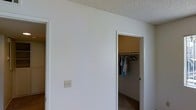 This single-story space presents a clean, straightforward design characterized by smooth white walls and a textured popcorn ceiling. The image highlights a walk-in closet accessible through a standard doorway, offering dedicated storage with a hanging rod already in place — a practical feature for everyday organization. The closet interior features warm-toned walls, creating a subtle contrast to the bright white of the main room. A standard light switch plate is mounted on the wall, and the flooring appears to be a neutral-toned hard surface, contributing to the space's understated, functional aesthetic. The open doorway to the left hints at additional rooms beyond, suggesting a connected layout throughout the single floor. Overall, the space maintains a simple, utilitarian character with minimal ornamentation, making it adaptable to a variety of uses and personal styles.