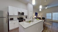 This 1-bedroom, 1-bathroom unit offers a thoughtfully designed single-floor layout that maximizes its compact footprint. The open-concept kitchen and living area serves as the heart of the home, featuring soaring ceilings that amplify the sense of space. The kitchen is outfitted with crisp white shaker cabinetry, granite countertops with a subway tile backsplash, a center island with a built-in sink, and a full suite of stainless steel appliances including a refrigerator, range, and over-the-range microwave. Warm wood-look flooring runs throughout the main living area, complementing the neutral tones of the walls.

The living space is bright and airy, with a front entry door flanked by large windows fitted with white blinds that allow for natural light control. The bedroom offers a cozy carpeted retreat with dual windows, providing ample natural light and a calm atmosphere.

The bathroom continues the home's consistent aesthetic, showcasing granite countertops, white cabinetry, a large vanity mirror, and a glass-enclosed shower, all set on wood-look flooring. Throughout the unit, high ceilings and a clean, modern design palette create an inviting and functional living environment.