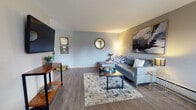 This single-floor residence in Mount Prospect, Illinois, offers a thoughtfully designed layout that balances comfort and functionality. The living room serves as the centerpiece of the space, featuring warm wood-look flooring, soft gray walls, and a baseboard heating system. A large sectional sofa anchors the room alongside a bold abstract canvas in a gold frame, a round decorative mirror, and a media console. Natural light filters in through a window at the far end of the room, where a dedicated workspace with a desk and seating provides a practical nook for everyday use.

The kitchen is well-appointed with white shaker-style cabinetry, gray composite countertops, a double-basin sink positioned beneath a window with an outdoor view, and a black range. The clean, neutral palette gives the kitchen a bright and organized feel.

The bedroom features plush carpeting, a vibrant teal accent wall, and a window with natural light. Two sets of bi-fold closet doors reveal generous hanging and shelving storage, making this room both stylish and practical.

The bathroom includes a full tub-shower combination with white tile surround, wood-look flooring, a single vanity with sink, and a small window that allows natural light into the space.
