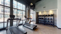 Located at 205 S High St in the heart of downtown Columbus, Ohio, this fitness facility combines industrial design with a high-performance training environment. The single-floor space is defined by soaring ceilings with exposed ductwork, structural concrete columns, and polished concrete floors that lend a bold, urban aesthetic throughout.

The main training floor is generously open, providing ample room for functional fitness, group workouts, and free movement. A motivational "One Hour for the Other 23" wall graphic anchors the central training zone, reinforcing the facility's performance-driven philosophy. Concept2 rowing machines, plyo boxes, kettlebells, and medicine ball racks are thoughtfully arranged across the space.

A dedicated cardio area features a row of treadmills positioned directly in front of floor-to-ceiling windows, offering views of the surrounding downtown streetscape and filling the zone with natural light. A separate strength training area houses a selection of cable and resistance machines, also bathed in natural light from large industrial-framed windows.

Near the facility's entrance, a reception and welcome area features a stylish counter, greenery, and cubbies for member use. A hydration station and display screen serve as a central amenity hub. Large ceiling fans and pendant lighting complete the well-equipped, modern training environment.