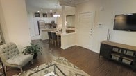 This single-floor space showcases an open-concept layout that seamlessly connects the living, dining, and kitchen areas into one cohesive, functional environment. Dark hardwood flooring runs throughout, providing a rich contrast against the warm cream and beige wall tones. Decorative columns with white trim add architectural interest while subtly defining the transition between spaces.

The kitchen is well-appointed with white cabinetry, stainless steel appliances including a full-size refrigerator and microwave, and a central island with a light stone countertop. Recessed lighting and a pendant chandelier illuminate the space effectively. An adjacent dining area accommodates a wooden table with upholstered chairs, positioned beneath a classic chandelier.

The living area features a glass-topped coffee table, a patterned area rug, and a decorative indoor plant that adds a natural touch. A solid white entry door with panel detailing and a visible thermostat panel are positioned near the entryway. Crown molding and clean baseboards throughout contribute to the polished, well-maintained character of the space.