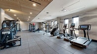 Located at 220 E Cimarron St in Colorado Springs, CO, this contemporary fitness center offers a well-appointed, single-floor workout space designed to accommodate a wide range of training needs. The facility is thoughtfully laid out across a generously sized open floor plan, finished with durable rubber tile flooring ideal for high-traffic exercise use.

The space features a distinctive slatted wood ceiling that adds warmth and visual interest, complemented by recessed lighting that ensures even illumination throughout. A mural of mountain silhouettes lines the walls, nodding to the Colorado landscape and creating an inspiring workout atmosphere. Large windows along one side of the facility allow natural light to pour in while framing views of the surrounding area.

Equipment is organized into dedicated zones. The cardio section is lined with treadmills, ellipticals, stair climbers, and stationary bikes, several equipped with individual screens for interactive training. The strength training area includes a multi-station cable and resistance machine setup, a power rack, and a well-stocked free weights section with medicine balls, kettlebells, and stability balls on organized storage racks. A flat-screen TV is mounted near a light wood cabinetry unit, offering additional entertainment or instructional viewing. The space also includes a mirrored wall and glass-partitioned areas, enhancing both functionality and the sense of openness.