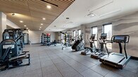 Located at 220 E Cimarron St in Colorado Springs, CO, this contemporary fitness center offers a well-appointed, single-floor workout space designed to accommodate a wide range of training needs. The facility is thoughtfully laid out across a generously sized open floor plan, finished with durable rubber tile flooring ideal for high-traffic exercise use.

The space features a distinctive slatted wood ceiling that adds warmth and visual interest, complemented by recessed lighting that ensures even illumination throughout. A mural of mountain silhouettes lines the walls, nodding to the Colorado landscape and creating an inspiring workout atmosphere. Large windows along one side of the facility allow natural light to pour in while framing views of the surrounding area.

Equipment is organized into dedicated zones. The cardio section is lined with treadmills, ellipticals, stair climbers, and stationary bikes, several equipped with individual screens for interactive training. The strength training area includes a multi-station cable and resistance machine setup, a power rack, and a well-stocked free weights section with medicine balls, kettlebells, and stability balls on organized storage racks. A flat-screen TV is mounted near a light wood cabinetry unit, offering additional entertainment or instructional viewing. The space also includes a mirrored wall and glass-partitioned areas, enhancing both functionality and the sense of openness.