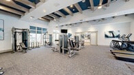 Located at 1001 NE Baker Rd in Stuart, Florida, this well-appointed fitness center offers a comprehensive, single-floor layout designed to support a full range of workout routines. The main gym area is expansive and airy, characterized by soaring ceilings with striking navy blue coffered beam detailing set against crisp white walls — a design element that lends the space a modern, upscale character. Ceiling fans are positioned throughout, ensuring comfortable air circulation during workouts.

The strength training zone is extensively equipped with a variety of cable machines, selectorized resistance equipment, a Smith machine with free weights, and additional functional training apparatus. Full-length mirrors line sections of the walls, enhancing the sense of space and allowing for proper form checks. A mounted flat-screen television provides entertainment in the main workout area.

A dedicated cardio section features treadmills and elliptical machines, positioned near large windows that frame views of the outdoor pool area, filling the zone with natural light.

A separate studio room, accessed through a sliding glass door, offers hardwood flooring, floor-to-ceiling mirrors, and expansive windows overlooking lush landscaping — ideal for yoga, stretching, or group fitness classes. The overall ambiance is bright, clean, and modern throughout.
