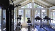 This fitness center is set within a single-story structure that combines a functional workout environment with an inviting, light-filled atmosphere. The space is defined by its striking vaulted ceiling with exposed white-painted beam detailing, lending a sense of height and openness that is rarely found in typical gym settings. Large multi-pane windows line the walls, stretching upward toward the peak of the roofline and flooding the interior with abundant natural light throughout the day. These expansive windows also frame picturesque views of surrounding mature trees and greenery, creating a refreshing outdoor connection during workouts. The interior walls feature a classic shiplap-style finish in a soft neutral tone, contributing to the space's cottage-like aesthetic while maintaining a clean and welcoming feel. The gym is equipped with a variety of cardio and strength training machines, including treadmills, an elliptical trainer, a stationary bike, and a cable weight machine, catering to a well-rounded fitness routine. The flooring provides a practical surface suited for exercise equipment. The overall ambiance strikes a balance between a bright, nature-connected environment and a purposefully outfitted fitness space, making it an appealing and motivating place to exercise.