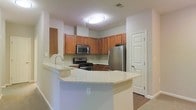This single-story space showcases a thoughtfully designed kitchen as its centerpiece. The kitchen features warm-toned wood cabinetry arranged in an L-shaped configuration, providing generous storage across both upper and lower cabinets. A full suite of stainless steel appliances includes a gas range, built-in microwave, and a French door refrigerator, offering a modern and functional cooking environment. Light granite countertops with subtle veining extend across the workspace and continue onto a peninsula that serves as a natural dividing element between the kitchen and the adjacent living area. The peninsula provides additional prep space and could accommodate seating for casual dining. The neutral light gray walls complement the warm cabinetry tones, creating a balanced and inviting atmosphere. Hardwood-style flooring is visible near the entryway, adding a touch of warmth to the overall aesthetic. Recessed flush-mount ceiling lighting ensures the kitchen remains well-lit throughout. A door adjacent to the refrigerator provides convenient access to an adjoining room or utility area, adding to the functional layout of the space.
