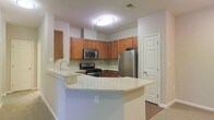 This single-story space showcases a thoughtfully designed kitchen as its centerpiece. The kitchen features warm-toned wood cabinetry arranged in an L-shaped configuration, providing generous storage across both upper and lower cabinets. A full suite of stainless steel appliances includes a gas range, built-in microwave, and a French door refrigerator, offering a modern and functional cooking environment. Light granite countertops with subtle veining extend across the workspace and continue onto a peninsula that serves as a natural dividing element between the kitchen and the adjacent living area. The peninsula provides additional prep space and could accommodate seating for casual dining. The neutral light gray walls complement the warm cabinetry tones, creating a balanced and inviting atmosphere. Hardwood-style flooring is visible near the entryway, adding a touch of warmth to the overall aesthetic. Recessed flush-mount ceiling lighting ensures the kitchen remains well-lit throughout. A door adjacent to the refrigerator provides convenient access to an adjoining room or utility area, adding to the functional layout of the space.