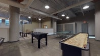 This single-floor recreational space is designed as a dedicated game room, offering a generous open layout that accommodates a diverse range of activities. The industrial-style architecture is defined by exposed ductwork, structural columns, and high ceilings with pendant and recessed lighting that illuminate the space effectively. Flooring throughout consists of patterned carpet tiles in neutral tones, contributing to a clean, functional aesthetic.

The main area houses several game tables arranged to allow comfortable circulation between each station. A foosball table serves as a central fixture, complemented by a full-size ping-pong table and a shuffleboard table positioned along one side. A dedicated billiards zone features a full-size pool table with overhead pendant lighting, a wall-mounted television, a cue rack, and a secondary foosball table, creating a well-appointed gaming alcove.

A reception or service counter is visible along one wall, suggesting a managed or monitored amenity space. Additional seating is available in a lounge area near the windows, which provide natural light to portions of the room. Multiple gray utility doors provide access to adjacent corridors, and clearly marked exit signage ensures safety throughout the space. Overall, the room presents a well-equipped, functional recreational environment suited for community or shared-use settings.