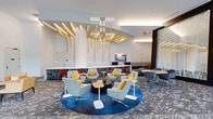 Located at 4 Vermella Way in Union, New Jersey, this generously proportioned single-floor amenity space makes an immediate impression with its soaring ceilings and thoughtfully curated interior design. The space unfolds as a series of distinct yet interconnected zones, each contributing to an energetic and creative atmosphere.

The main lounge area anchors the space with a striking sculptural ceiling installation of gold-toned wooden fins and cascading pendant lighting, suspended above a circular blue area rug surrounded by plush striped swivel chairs with bold yellow accent pillows. Nearby, a bar-height counter with red seating and a large display screen provides a functional social hub near the glass entrance.

Adjacent areas feature dramatic double-height ceilings adorned with geometric hanging sculptures in green, blue, and white, floating above modular lounge seating arranged around colorful wall-mounted artwork. Glass-walled private meeting rooms with black steel framing line one corridor, offering transparency while maintaining acoustic separation.

A vibrant mural reading "Creativity Is In All Of Us" commands one wall, bathed in natural light from floor-to-ceiling windows with outdoor views. Hallway transitions are accented with motivational typographic artwork and warm wood-paneled walls. Throughout, patterned carpet, curated plants, and layered lighting unify the space into a cohesive, inspiring environment.