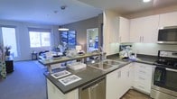 This single-floor residence showcases a thoughtfully designed open-concept layout that connects the kitchen, dining, and living areas into one cohesive space. The kitchen is a standout feature, equipped with white cabinetry, dark countertops, a stainless steel double sink, and a built-in dishwasher. A peninsula with a breakfast bar extends into the dining area, offering both additional workspace and casual seating. Pendant lighting overhead adds a modern, polished touch. The backsplash features decorative glass tiles, contributing to the kitchen's contemporary aesthetic. The living area, visible from the kitchen, is well-lit by a large window that invites natural light into the space. Gray accent walls contrast cleanly against white trim and light-toned furnishings, lending the interior a calm, modern atmosphere. A glimpse into what appears to be a bedroom reveals a similarly styled space with coordinated décor. The combination of dark hardwood-style flooring, neutral carpeting, and modern light fixtures throughout creates a unified, stylish environment suited for comfortable everyday living.