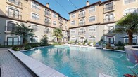 Located in the vibrant Uptown neighborhood of Dallas, Texas, this beautifully designed courtyard amenity space showcases a Mediterranean-inspired architectural style that sets a refined and welcoming tone throughout. The centerpiece of the space is a generously sized resort-style swimming pool with clear blue water, complemented by a decorative fountain that adds a touch of elegance to the atmosphere. Surrounding the pool, a spacious cobblestone patio provides ample room for relaxation and socializing, lined with a row of sleek chaise lounge chairs ideal for soaking up the Texas sun. Mature palm trees and carefully manicured shrubbery add lush, tropical character to the courtyard, while string lights draped overhead create a warm, inviting ambiance during evening hours. An outdoor grilling station with a built-in gas grill offers a convenient space for al fresco dining and entertaining. The multi-story building façade features arched windows, wrought iron balcony railings, and a warm stucco exterior that frames the courtyard on all sides, creating a private and enclosed retreat. Paved walkways with neatly trimmed hedgerows connect the various areas of the courtyard, and classic lantern-style light fixtures enhance the space's charming aesthetic. This thoughtfully designed outdoor space delivers both functionality and visual appeal.