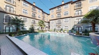 Located in the vibrant Uptown neighborhood of Dallas, Texas, this beautifully designed courtyard amenity space showcases a Mediterranean-inspired architectural style that sets a refined and welcoming tone throughout. The centerpiece of the space is a generously sized resort-style swimming pool with clear blue water, complemented by a decorative fountain that adds a touch of elegance to the atmosphere. Surrounding the pool, a spacious cobblestone patio provides ample room for relaxation and socializing, lined with a row of sleek chaise lounge chairs ideal for soaking up the Texas sun. Mature palm trees and carefully manicured shrubbery add lush, tropical character to the courtyard, while string lights draped overhead create a warm, inviting ambiance during evening hours. An outdoor grilling station with a built-in gas grill offers a convenient space for al fresco dining and entertaining. The multi-story building façade features arched windows, wrought iron balcony railings, and a warm stucco exterior that frames the courtyard on all sides, creating a private and enclosed retreat. Paved walkways with neatly trimmed hedgerows connect the various areas of the courtyard, and classic lantern-style light fixtures enhance the space's charming aesthetic. This thoughtfully designed outdoor space delivers both functionality and visual appeal.