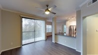 This single-floor residence showcases an open-concept design that seamlessly connects the living area and kitchen, creating a spacious and functional layout. The living area features smooth hardwood-style flooring throughout, complemented by crown molding along the ceiling and a ceiling fan with a built-in light fixture that enhances both comfort and ambiance. Large sliding glass doors lined with vertical blinds flood the space with abundant natural light while offering views of lush greenery outdoors, creating a bright and airy atmosphere.

The kitchen is positioned at the rear of the open floor plan and is separated by a structural support column, lending subtle definition to the space. It is equipped with stainless steel appliances, including a full-size refrigerator, and features dark-toned countertops atop a peninsula-style breakfast bar with a built-in sink. White cabinetry lines the upper walls, and recessed track lighting overhead highlights the kitchen's clean, modern aesthetic. An additional window in the kitchen area allows for extra natural light, further enhancing the bright, welcoming character of the overall space.