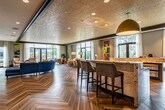 This elegantly designed single-floor community lounge is a standout gathering space that masterfully blends contemporary style with inviting comfort. The defining feature of the room is its striking herringbone-patterned wood flooring, which flows seamlessly throughout and anchors the entire space with warmth and visual interest.

At the heart of the room sits a curved, circular bar island topped with richly veined granite countertops and surrounded by upholstered bar stools. Beneath the countertop, integrated appliance drawers and cabinetry add functional elegance, while a large pendant light in a bold gold finish hangs overhead, drawing the eye upward to the textured ceiling with recessed lighting.

Multiple distinct seating zones are thoughtfully arranged throughout the space, featuring a mix of plush curved sofas in deep navy velvet, rounded lounge chairs in warm neutral tones, and accent chairs with mustard yellow throw pillows — all contributing to a vibrant yet cohesive color palette. Colorful abstract artwork adorns the walls, reinforcing the contemporary aesthetic.

Expansive floor-to-ceiling windows and glass-paneled double doors flood the interior with natural light and provide direct access to an outdoor terrace. A media wall with a mounted flat-screen television adds an entertainment-focused zone to the space. The overall ambiance is refined, sociable, and thoughtfully curated for both relaxation and community engagement.