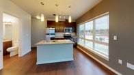 Located on the fourth floor at 1149 NW Market Street in Seattle's Ballard neighborhood, this unit features a thoughtfully designed open-concept layout finished with rich hardwood flooring throughout the main living areas. The kitchen serves as a central focal point, anchored by a granite-topped island with a double stainless steel sink and pendant lighting overhead. Dark-stained cabinetry extends to the ceiling, providing generous storage, while stainless steel appliances—including a refrigerator, range, and microwave—complete the functional and polished aesthetic. Track lighting and a window overlooking the balcony bring additional natural light into the cooking space.

The living area is bright and open, benefiting from large sliding glass doors that lead to a private balcony with views of the surrounding urban streetscape. The bedroom features plush carpeting and a sliding-door closet with built-in shelving, offering practical storage solutions. The bathroom is well-appointed with a granite-topped vanity with dark wood cabinetry, a large mirror, a glass-enclosed walk-in shower, and wood-look flooring. The overall design balances contemporary finishes with a clean, neutral palette, creating a cohesive and welcoming living environment.