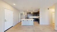 This single-floor residence offers four bedrooms and two bathrooms thoughtfully arranged along a central hallway. The kitchen serves as a functional centerpiece, featuring rich dark espresso cabinetry reaching toward the ceiling, white quartz countertops, stainless steel appliances including a refrigerator, range, and microwave, and a spacious island with bar seating illuminated by pendant lighting. Light wood-style flooring flows throughout the main living and kitchen areas, creating a cohesive and modern aesthetic. The open layout connects the kitchen to a generously sized living area that benefits from a wide bank of windows, filling the space with natural light and providing outdoor views. The four bedrooms are comfortably sized, each featuring plush carpeting and windows with blinds for privacy and natural light. The primary bedroom includes access to a dedicated walk-in closet equipped with wire shelving and hanging rods on multiple walls for ample storage. One bathroom conveniently houses a full-size washer and dryer alongside a vanity with white countertop and a large mirror, combining laundry and bath functions efficiently. Neutral warm-toned walls throughout the residence create a calm and inviting ambiance, while recessed lighting and clean white trim contribute to the overall contemporary feel of this well-designed home.