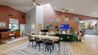 Located in Roanoke, Virginia, this single-floor community clubhouse on Glade Creek Blvd NE offers a thoughtfully designed gathering space with a warm, modern aesthetic. The centerpiece is a dramatic great room boasting soaring vaulted ceilings accentuated by clerestory windows that flood the space with natural light. Boldly painted terracotta and mauve accent walls provide a rich backdrop for the open-plan layout, which features a large communal dining table, a wall-mounted flat-screen TV, and vibrant green lounge seating—perfect for social gatherings. Ceiling fans throughout ensure comfort year-round, while light hardwood flooring ties the areas together seamlessly. Adjacent to the great room, a cozy lounge area offers a fireplace, plush swivel armchairs, and large windows with floor-length drapes, creating an inviting retreat. A dedicated office area with multiple desks and ample natural light from oversized windows provides a functional workspace. The kitchen is well-appointed with warm wood cabinetry, dark granite-style countertops, a stainless steel refrigerator, and a convenient exterior door with outdoor views. This versatile, generously scaled space is designed to serve as a true community hub.