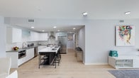 This single-floor space showcases a thoughtfully designed open-concept kitchen that serves as the centerpiece of the layout. The kitchen is outfitted with crisp white flat-panel cabinetry, stainless steel appliances including a French door refrigerator and dishwasher, and a stainless steel wall-mounted range hood above the cooktop. A contrasting dark-framed kitchen island with a bright white countertop provides additional workspace and casual seating with black bar stools, adding visual interest against the otherwise light palette. Recessed lighting is distributed evenly across the white ceiling, ensuring the space is well-lit throughout. Light-toned hardwood flooring runs continuously through the kitchen and into the adjoining hallway, creating a cohesive and airy feel. The neutral gray-white walls contribute to the clean, modern aesthetic. A corridor extending from the kitchen leads to additional rooms, suggesting a practical and connected floor plan. The overall ambiance is contemporary and functional, with a minimalist design sensibility that emphasizes clean lines and a bright, open atmosphere.