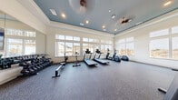 This fitness center is a thoughtfully designed, single-floor workout facility offering a generous open floor plan ideal for a full range of exercise activities. The space is defined by its striking tray ceiling, finished in a cool slate blue and outfitted with recessed lighting and dual ceiling fans, creating a polished and comfortable atmosphere. Durable speckled rubber flooring runs throughout, providing excellent traction and cushioning underfoot.

Large, multi-pane windows line multiple walls, flooding the interior with natural light and offering pleasant views of the surrounding outdoor landscape, including a pergola and open sky. The abundance of glazing contributes to an airy, energizing ambiance well-suited to physical activity.

The cardio zone is equipped with treadmills, elliptical machines, and a stationary spin bike, all positioned along the window-facing wall to take advantage of the outdoor views. The strength training area features a multi-function cable and weight machine, a dumbbell rack with a full range of free weights, and ample open floor space for functional training.

Full-length wall mirrors span a significant portion of the interior, visually expanding the space and serving a practical purpose for form monitoring during workouts. A wall-mounted flat-screen television is also present. A separate entry area provides a transitional space with posted fitness center guidelines, completing this well-appointed community amenity.