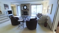 This two-floor home offers a comfortable and well-appointed living environment designed for both relaxation and everyday functionality. The living room serves as the heart of the space, anchored by a striking floor-to-ceiling brick fireplace with a wood mantel and decorative mirror, creating a classic and cozy focal point. A large sliding glass door floods the room with natural light and provides direct access to an outdoor patio area, where garden views and outdoor seating can be enjoyed. The living area is generously sized, accommodating a full seating arrangement including plush swivel chairs and a sofa grouped around a glass-top coffee table with ornate wrought-iron detailing. Soft, neutral carpet flows throughout the main living space, contributing to the warm and welcoming ambiance. An additional doorway provides a view into adjoining spaces, hinting at the home's thoughtful layout and connectivity between rooms. The overall design blends traditional elements — such as the brick fireplace and classic furnishings — with practical features like ample natural light and indoor-outdoor flow. Spread across two floors, this home offers distinct living zones suited to a variety of lifestyles and needs.