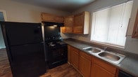 This single-story unit at University Lofts offers a practical and straightforward layout designed to accommodate everyday living. The kitchen is equipped with warm oak cabinetry — both upper and lower — dark laminate countertops, a double-basin stainless steel sink, a black electric range with overhead ventilation, and a full-size refrigerator. A window with blinds allows natural light into the cooking space, and wood-look vinyl plank flooring runs throughout the kitchen and adjoining areas.

The living and entry areas also feature durable wood-look flooring, providing a cohesive look through the main gathering spaces. A front entry door is flanked by a window, allowing additional natural light into the home. The bedrooms are carpeted and finished with neutral gray walls and white trim. Several bedrooms include windows with blinds that offer views of surrounding greenery. One bedroom features a dedicated closet with a built-in rod and shelving, along with access to additional doors for storage or utility purposes.

The full bathroom includes a single-sink vanity with wood cabinetry and laminate counters, a toilet, and a full tub-shower combination with a white surround. Light tile flooring completes the space, keeping the overall aesthetic clean and functional.
