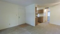 This single-floor residence offers a straightforward and functional layout designed for comfortable everyday living. The main living area features low-pile carpeting that extends throughout, providing a warm and practical foundation underfoot. A partial wall divides the living space from the kitchen, creating a sense of separation while maintaining an open feel between the two areas.

The kitchen is efficiently arranged with medium-toned wood cabinetry on both the upper and lower sections, offering ample storage. A laminate countertop runs along the base cabinets, and the kitchen floor transitions to a hard surface, distinguishing it from the carpeted living space. A built-in appliance space is visible beneath the counter, adding to the kitchen's utility.

The entryway, located adjacent to the partial dividing wall, features a solid white door with a deadbolt, opening directly into the living area. The ceilings have a subtle texture finish, and the walls are painted in a neutral, light tone throughout, contributing to a clean and understated aesthetic. A hallway visible beyond the kitchen suggests additional rooms, with overhead lighting providing consistent illumination throughout the space. Overall, the residence presents a practical and well-organized single-level floor plan.