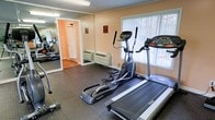 This fitness room offers a focused and functional workout environment designed to meet essential exercise needs. The space is outfitted with key cardio equipment, including a treadmill and an elliptical trainer, both positioned to take advantage of the natural light streaming in through large windows that look out onto a lush, tree-lined exterior. The flooring features durable, textured rubber matting, providing stability and safety during workouts. A full-length wall mirror along one side of the room visually expands the space while allowing users to monitor their form during exercise. The walls are finished in a warm, neutral tone, contributing to an inviting and energizing atmosphere. A wall-mounted HVAC unit ensures year-round climate comfort within the room. Additional free weights are visible near the mirrored wall, offering supplementary strength training options. The room is well-lit with overhead fluorescent lighting, ensuring visibility at any hour. A single entry door with an adjacent notice board provides easy access and keeps users informed of any facility guidelines. Overall, this fitness room is a practical and well-appointed space suited for daily cardio and light strength training routines.