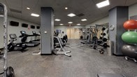 This fitness center is a fully outfitted, single-level gym designed to accommodate a wide variety of training needs. The space features a drop ceiling with recessed panel lighting that provides consistent, even illumination throughout the floor plan. Durable, patterned modular carpet tiles cover the entire floor, offering both comfort and practicality underfoot.

The cardio zone is equipped with treadmills, elliptical trainers, and upright stationary bikes, several of which are mounted with personal viewing screens. A dedicated strength training area houses a multi-station cable and pulley machine as a centerpiece, complemented by Smith machines, barbell racks, and multiple weight benches for compound lifts. A full-length wall mirror anchors the free weights section, where a complete rack of dumbbells is neatly organized alongside BOSU balance trainers and a flat utility bench.

Structural columns punctuate the open floor plan, lending an industrial character while subtly defining workout zones. The entrance features frosted double glass doors, and informational bulletin boards are posted near the entry area. A fire extinguisher and emergency exit signage reflect strong safety compliance throughout. The neutral gray and white color palette, paired with motivational fitness artwork on the walls, creates a focused and professional training environment suitable for all fitness levels.