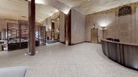The lobby at 544 Broad Street in Newark, New Jersey is a masterfully preserved Art Deco space that blends historic grandeur with thoughtful modern touches. Floor-to-ceiling marble wall panels in warm rose and deep burgundy tones set an authoritative architectural tone throughout, while expansive terrazzo flooring with subtle geometric patterning anchors the sweeping open layout. Intricate gilded friezes and ornamental gold detailing line the upper walls and ceiling borders, drawing the eye upward to dramatized ceiling planes adorned with geometric relief work. Brass torch-style sconces and freestanding floor lamps cast a warm, ambient glow that highlights the richness of the original materials. Elevator banks clad in brushed gold metalwork punctuate the space at measured intervals, maintaining the period aesthetic. A dedicated mail room features sleek black mailbox banks lining three walls, a sculptural pendant light, and slate-toned tile flooring. Throughout the main lobby, carefully curated contemporary seating—including velvet sofas, lounge chairs, and marble-topped side tables—provides comfortable gathering areas without compromising the historic character. An ornamental memorial plaque serves as a respectful focal point along one corridor wall. The overall atmosphere is one of distinguished civic elegance, honoring the building's storied past while remaining fully functional for today's occupants.