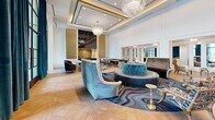 Located at 4343 Woodhead St in Houston's Montrose neighborhood, this single-floor amenity space delivers a refined, hotel-inspired aesthetic across a generously proportioned layout. The main living areas are defined by soaring ceilings, intricate coffered details trimmed in gold, and large arched floor-to-ceiling windows dressed in rich teal velvet drapes that flood the interior with natural light and offer views to the outdoors. Herringbone-patterned hardwood flooring flows throughout, complemented by elegant crystal chandeliers and gold-accented wall sconces. The primary lounge showcases a curvaceous sectional sofa, tufted ottomans, and sculptural accent chairs arranged around a swirling blue-patterned area rug, while a secondary lounge area provides additional intimate seating with marble-topped tables and mirrored accents. A connecting hallway features marble tile flooring with dark border inlays and crisp white millwork, maintaining the space's polished character. The private screening room offers a dramatic contrast, enveloped in dark tones with a coffered ceiling trimmed in warm wood, deep charcoal velvet curtains, a large projection screen, and plush oversized lounge chairs arranged on tiered, patterned carpet for an immersive cinematic experience.