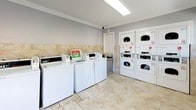 This shared laundry facility is thoughtfully designed to offer both functionality and comfort. The room is finished with elegant diagonal-set travertine-style tile wainscoting that lines the lower walls, complemented by matching tile flooring that gives the space a clean, polished appearance throughout.

The laundry area is equipped with a row of top-loading washing machines and stacked dryer units, providing ample capacity for residents. Each machine features coin-operated controls, and a waste receptacle is conveniently positioned nearby for ease of use.

A bold red entry door with glass panel detailing serves as a welcoming focal point, providing direct access from the exterior. Large windows allow generous natural light to fill the room, creating a bright and airy atmosphere. The seating area, furnished with a wooden bench, offers a comfortable place to wait during laundry cycles.

Crown molding adds an architectural detail to the ceiling, elevating the overall finish of the space beyond a typical utility room. Overhead fluorescent lighting ensures the room remains well-lit at all times. A security camera is also visible, providing an added sense of safety for users. The layout is open and uncluttered, making navigation easy and the overall laundry experience efficient and pleasant.
