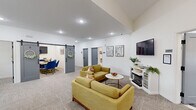 This thoughtfully designed single-floor space blends comfort and functionality across a well-appointed open layout. The main lounge area features plush gold-toned sofas arranged around a marble-top coffee table, a wall-mounted flat-screen TV, and an electric fireplace set within a crisp white media console — creating a warm, inviting atmosphere for casual gatherings or downtime. Recessed lighting and elevated ceilings enhance the sense of openness throughout.

A dedicated work zone includes a workstation with dual monitors and ergonomic seating, positioned beneath a window that allows natural light to filter in. The space is branded with a marquee "ENCLAVE" sign, reinforcing its identity as a curated community hub. A stylish navy accent wall anchors the coffee bar area, complete with white cabinetry, a sink, and a statement sarburst chandelier overhead — ideal for residents or members to enjoy a beverage before settling in.

A charming sliding barn door leads to a private conference or meeting room outfitted with blue task chairs and a collaborative table. The flooring transitions between neutral patterned carpet and decorative tile, delineating functional zones while maintaining a cohesive aesthetic. Throughout, clean white walls, modern fixtures, and thoughtful décor create a polished yet welcoming environment.
