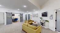This thoughtfully designed single-floor space blends comfort and functionality across a well-appointed open layout. The main lounge area features plush gold-toned sofas arranged around a marble-top coffee table, a wall-mounted flat-screen TV, and an electric fireplace set within a crisp white media console — creating a warm, inviting atmosphere for casual gatherings or downtime. Recessed lighting and elevated ceilings enhance the sense of openness throughout.

A dedicated work zone includes a workstation with dual monitors and ergonomic seating, positioned beneath a window that allows natural light to filter in. The space is branded with a marquee "ENCLAVE" sign, reinforcing its identity as a curated community hub. A stylish navy accent wall anchors the coffee bar area, complete with white cabinetry, a sink, and a statement sarburst chandelier overhead — ideal for residents or members to enjoy a beverage before settling in.

A charming sliding barn door leads to a private conference or meeting room outfitted with blue task chairs and a collaborative table. The flooring transitions between neutral patterned carpet and decorative tile, delineating functional zones while maintaining a cohesive aesthetic. Throughout, clean white walls, modern fixtures, and thoughtful décor create a polished yet welcoming environment.