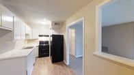This single-story residence at Fern features two bedrooms and one and a half bathrooms across a practical, well-organized floor plan. The kitchen is outfitted with white cabinetry, a black electric range, refrigerator, and dishwasher, all set against light wood-style flooring that extends through the main living areas. The neutral cream-toned walls throughout create a bright, clean atmosphere, while a textured ceiling adds subtle character. An open doorway from the kitchen provides a seamless transition into the carpeted bedroom areas, where soft flooring adds warmth and comfort underfoot. The layout promotes easy flow between spaces, with clearly defined zones for living and sleeping. Overhead ventilation is visible in the kitchen area, reflecting practical utility features integrated throughout the unit. The overall ambiance is clean, understated, and move-in ready, making it a straightforward and functional living space suited for a variety of occupants.
