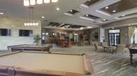 This expansive single-floor communal space is thoughtfully designed to accommodate a variety of social and recreational activities. The open-plan layout flows seamlessly from one zone to the next, creating a welcoming and versatile environment. A full-size billiards table anchors the foreground of the space, making it a natural focal point for recreation and entertainment. Beyond the billiards area, a dedicated dining section features a round table surrounded by richly upholstered burnt-orange chairs beneath a stylish chandelier, adding warmth and elegance to the space. Further into the room, a comfortable lounge area offers plush seating arranged for relaxed conversation. A striking stone fireplace surround serves as a decorative architectural centerpiece along one wall, complemented by additional media and display elements. The ceiling is a standout feature, showcasing coffered and tray details with dark wood beam accents and recessed lighting throughout, adding depth and visual interest. Large windows and glass doors allow natural light to filter into the space, enhancing its bright and airy ambiance. The neutral palette of grays and taupes, accentuated by warm orange furnishings and natural stone elements, creates a sophisticated yet inviting atmosphere suited for community gatherings and leisure activities.