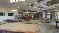 This expansive single-floor communal space is thoughtfully designed to accommodate a variety of social and recreational activities. The open-plan layout flows seamlessly from one zone to the next, creating a welcoming and versatile environment. A full-size billiards table anchors the foreground of the space, making it a natural focal point for recreation and entertainment. Beyond the billiards area, a dedicated dining section features a round table surrounded by richly upholstered burnt-orange chairs beneath a stylish chandelier, adding warmth and elegance to the space. Further into the room, a comfortable lounge area offers plush seating arranged for relaxed conversation. A striking stone fireplace surround serves as a decorative architectural centerpiece along one wall, complemented by additional media and display elements. The ceiling is a standout feature, showcasing coffered and tray details with dark wood beam accents and recessed lighting throughout, adding depth and visual interest. Large windows and glass doors allow natural light to filter into the space, enhancing its bright and airy ambiance. The neutral palette of grays and taupes, accentuated by warm orange furnishings and natural stone elements, creates a sophisticated yet inviting atmosphere suited for community gatherings and leisure activities.