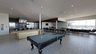 This contemporary amenity space is designed with a sleek, open-concept layout that effortlessly blends entertainment and social gathering areas. At the heart of the room sits a full-sized billiards table, serving as a central focal point beneath a bright, recessed-lit ceiling. The light-toned plank flooring runs throughout, lending a clean and cohesive aesthetic to the entire space.

Toward one side, a stylish bar area features a light wood counter surface and a dark stone backsplash, accented by industrial-style pendant lighting that adds a warm, modern touch. Adjacent to the bar, a dining and lounge zone is furnished with high-top tables and bar-height chairs, ideal for casual conversation or collaborative gatherings. A wall-mounted television is integrated into a richly paneled accent section, creating an entertainment nook that complements the overall design.

Large floor-to-ceiling windows along the perimeter flood the space with natural light and offer views to an outdoor area, seamlessly connecting the interior with the exterior environment. The high ceilings and open floor plan contribute to an airy, expansive feel. Overall, the space strikes a sophisticated balance between functionality and modern design, making it well-suited for both relaxed recreation and social events.