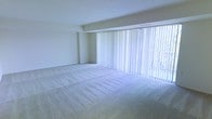 This single-floor space offers a clean, understated aesthetic defined by neutral tones and a sense of openness. The main living area is finished with wall-to-wall plush carpeting in a soft, light beige tone, providing a comfortable and cohesive foundation throughout the room. A defining architectural feature is the expansive floor-to-ceiling window wall spanning the width of the room, dressed with full-length vertical blinds that diffuse natural light evenly across the interior. When the blinds are drawn, glimpses of outdoor greenery are visible, reinforcing a connection to the outside environment. The walls are finished in a crisp white, complementing the bright, airy atmosphere created by the generous glazing. A subtle ceiling beam detail adds mild architectural interest without disrupting the clean lines of the space. The overall ambiance is calm and neutral, offering a versatile blank canvas well-suited for a variety of furnishing arrangements and interior styles. The single-floor layout ensures ease of movement and accessibility throughout.