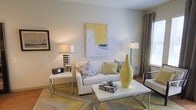 This single-floor space showcases a thoughtfully designed interior with a clean, modern aesthetic. The living area serves as the heart of the space, centered around a crisp white sofa adorned with patterned grey and green accent pillows. A sleek white coffee table with chrome legs anchors the seating area, styled with decorative accessories that add personality and charm. The color palette throughout is cohesive and inviting, blending soft whites and warm greys with pops of yellow — reflected in the statement vase, area rug, and abstract wall art that adds a sophisticated focal point above the sofa. Warm ambient lighting is provided by two stylish table and floor lamps, including one with a bold chevron-patterned base, creating a welcoming atmosphere. Neutral walls allow the carefully chosen décor elements to stand out, while draped curtains frame a window that introduces natural light into the room. The overall ambiance is contemporary yet comfortable, striking a balance between curated design and livable warmth. The space is well-appointed and move-in ready, offering a polished environment suited for both relaxing and entertaining.