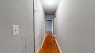This single-floor unit is characterized by rich, polished hardwood flooring that runs throughout the main living areas, hallways, and bedrooms, lending a warm, cohesive aesthetic against the cool grey-toned walls. A narrow hallway connects the rooms, with flush-mount ceiling lighting and an electrical panel access point along the wall.

The main living area is generously proportioned, featuring a ceiling fan and direct sightlines to both the kitchen and an additional room, creating an open, connected flow. Multiple windows admit abundant natural light throughout. Two bedrooms each feature wall-mounted ductless split A/C units, multiple windows offering outdoor views, and the same gleaming hardwood floors.

The kitchen is compact and functional, equipped with dark cabinetry, a black electric range, a full-size refrigerator, and a sink beneath a window. Wood-look vinyl flooring grounds the space. The bathroom features white subway tile wainscoting, a window for ventilation and natural light, a soaking tub, toilet, and vanity, with wood-look flooring tying together the practical, clean design.