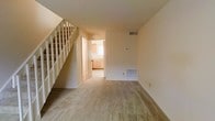 This two-story space offers a straightforward and functional layout designed for comfortable living. The main level features a generously sized living area finished with wall-to-wall carpet in a neutral tone, creating a warm and inviting atmosphere. Smooth, light-colored walls throughout contribute to a clean, airy feel. A white-balustered open staircase runs along one side of the living area, providing access to the upper floor while adding a classic architectural detail to the space. An HVAC floor vent and a wall-mounted thermostat are visible, indicating a dedicated climate control system. Through a doorway at the rear of the main level, the kitchen is accessible and features light wood cabinetry and a window that brings in natural light, brightening the space. The transition between the living area and kitchen is defined by a subtle shift in flooring. Overall, the space maintains a neutral, move-in-ready aesthetic with a cohesive color palette of beige and cream tones throughout, offering a versatile foundation for personalization across both levels.
