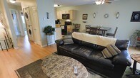 This single-floor space is designed around an open-concept layout that seamlessly connects the living room, kitchen, and dining area, creating a cohesive and functional environment. The living room serves as the central gathering point, anchored by a dark leather sofa accented with patterned throw pillows and complemented by a marble-style round coffee table atop a neutral area rug. Warm hardwood flooring flows throughout, adding a natural, inviting tone to the space.

The kitchen is positioned just beyond the living area, featuring light wood cabinetry, black appliances including a range and microwave, and a light-toned countertop that brightens the cooking space. A peninsula-style counter creates a natural visual boundary between the kitchen and living room while maintaining the open feel.

Adjacent to the kitchen, a dining area accommodates a dark wood table with matching chairs, ideal for everyday meals. Soft sage-green walls run throughout the space, contributing to a calm and cohesive atmosphere. A ceiling fan provides practical comfort, while a laundry area is conveniently tucked near the entrance hallway, adding to the overall functionality of this well-organized, single-level home.